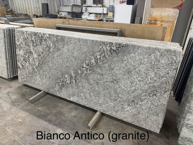 A large, rectangular slab of Bianco Antico granite with a gray and white speckled pattern is displayed upright in a warehouse. The text Bianco Antico (granite) appears at the bottom of the image.