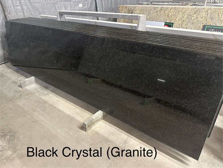 A polished slab of Black Crystal granite is standing upright in a warehouse, labeled Black Crystal (Granite) in black text at the bottom of the image.