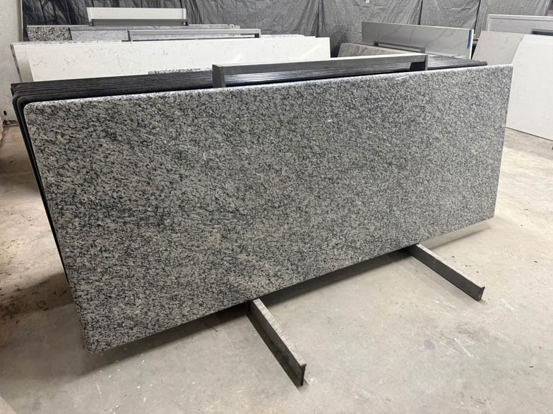 A large rectangular slab of polished gray granite with black and white speckles stands upright on a metal frame in a workshop with other stone slabs in the background.