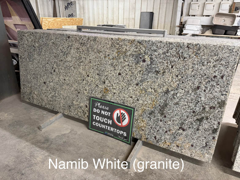 A large slab of Namib White granite stands upright in a warehouse, supported by metal stands. A sign in front reads Please do not touch countertops. Text at the bottom labels the granite as Namib White (granite).