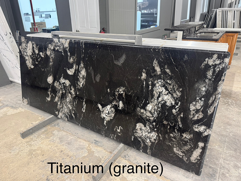 A large, polished slab of black granite with striking white and gray swirling patterns, labeled Titanium (granite). The slab is displayed upright in a showroom with other stone samples in the background.