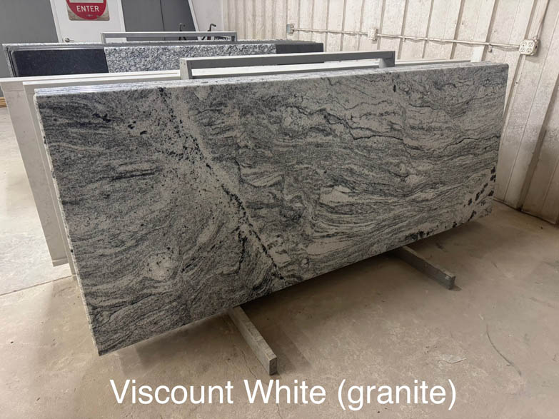 A large slab of Viscount White granite with black and gray swirling patterns stands on a metal rack in a warehouse. The text Viscount White (granite) is written at the bottom of the image.