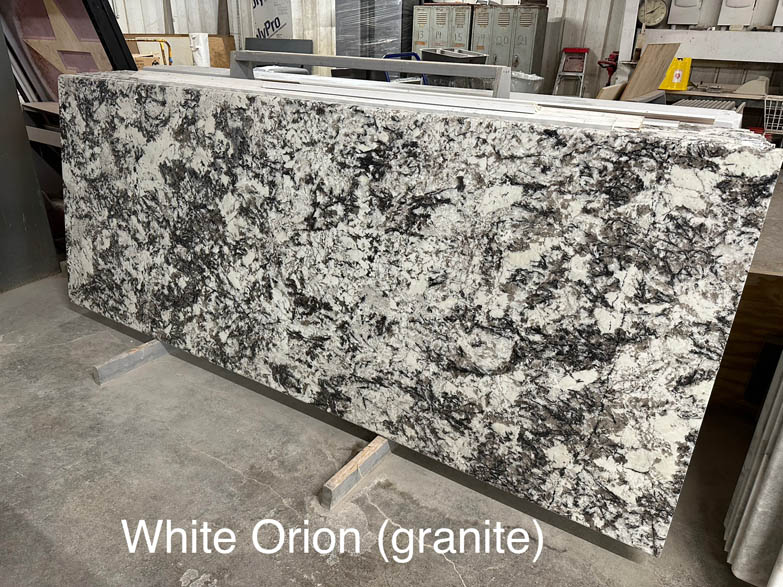 A large slab of White Orion granite with a white and black speckled pattern, displayed indoors on a stand. The text White Orion (granite) is overlaid at the bottom of the image.