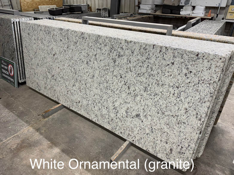 A large slab of White Ornamental granite with a predominantly white and gray speckled pattern is standing upright in a warehouse. The text White Ornamental (granite) appears at the bottom of the image.